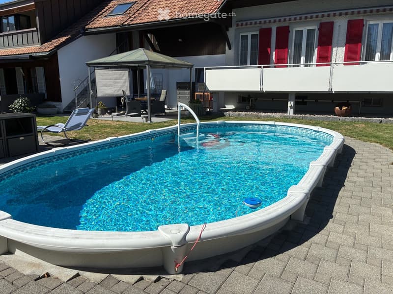 A private swimming pool with a modern design, surrounded by a paved patio and a well-maintained lawn. Nearby, there is a gazebo with outdoor furniture and a lounge chair.