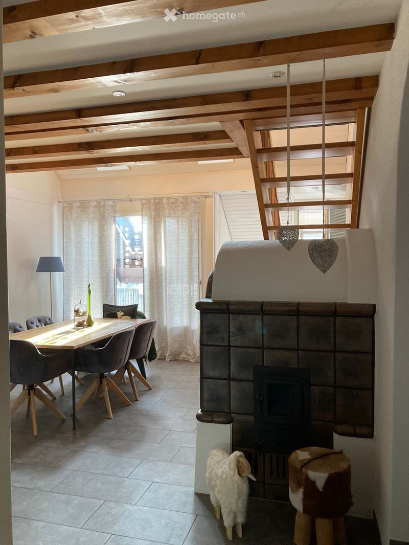 Dining area with a wooden table, chairs, and a fireplace with a decorative stool in front. There is a lamp, a vase on the table, and curtains on the windows.