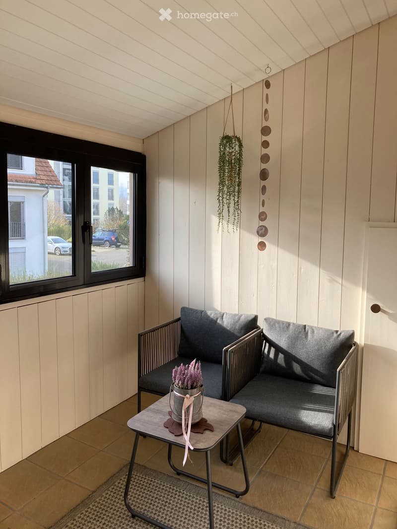 Cozy balcony with wooden walls, grey sofa, small table with a flower pot, hanging plant, and decorative wind chime.