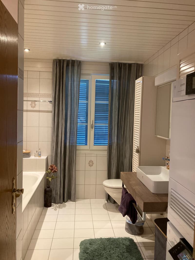Modern bathroom with white tiles, a bathtub, toilet, sink, and a large window with curtains. Includes a wooden vanity, a mirror, a decorative vase, and a green rug.