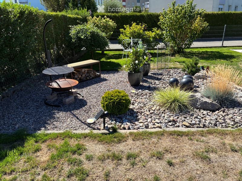 Well-maintained garden with gravel pathways, decorative plants, a fire pit, a swing, and a bench. Features include spherical sculptures, potted plants, and a stack of firewood.