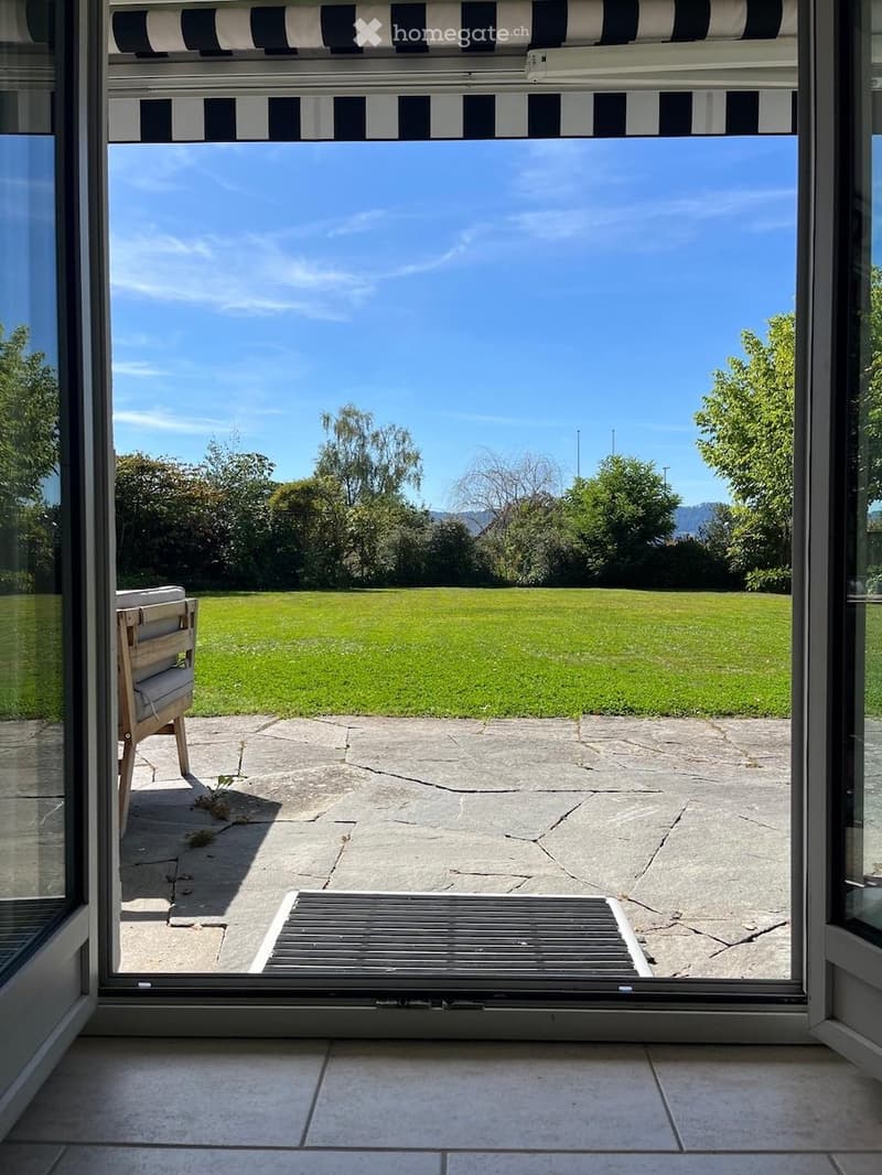 View from inside a house onto a well-maintained garden with a lawn, trees, and a clear blue sky. A wooden chair is visible on the patio.