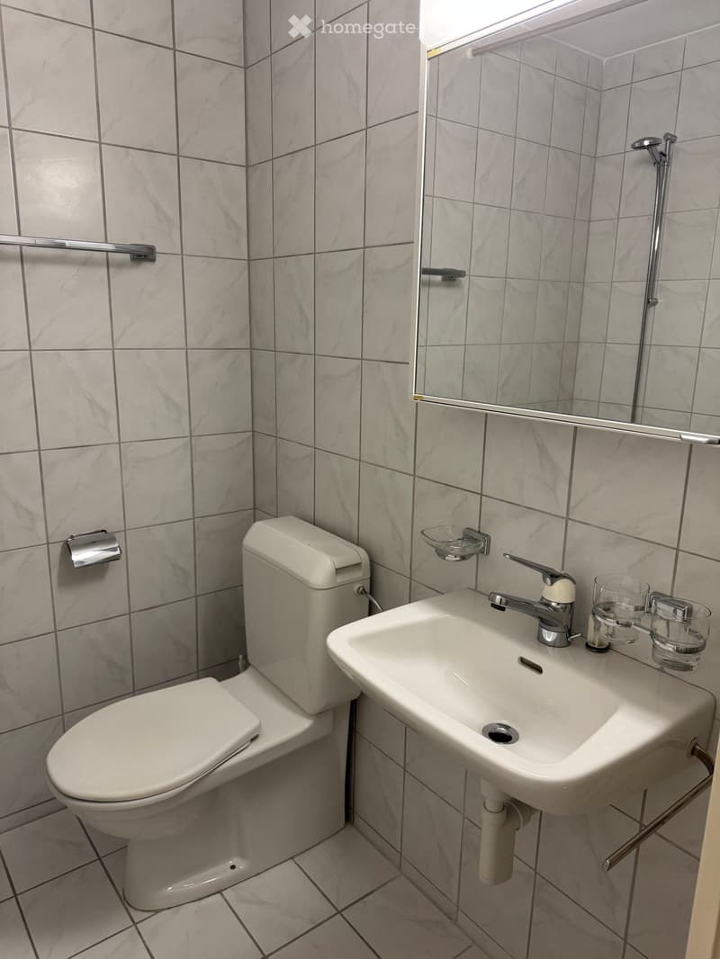 A small bathroom with a toilet, sink, and mirror. The walls are tiled in a light grey marble pattern, and the floor is tiled in a light grey pattern as well. There is a glass on the sink counter.