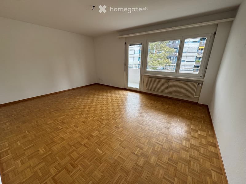 Empty room with wooden herringbone flooring, white walls, large windows, and a radiator under the windows.