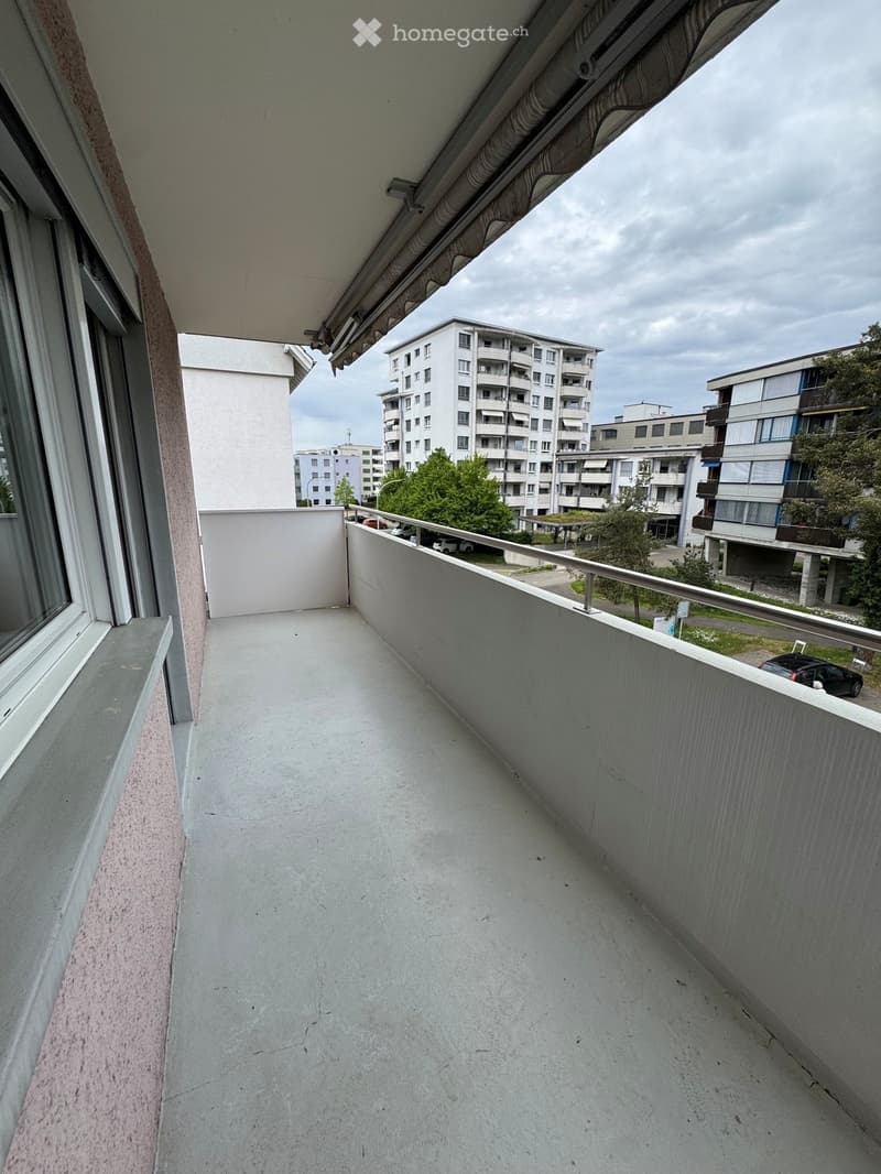 Empty balcony with concrete floor, metal railing, and a view of surrounding buildings and greenery.