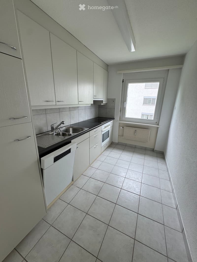 Modern kitchen with white cabinets, tiled floor, sink, oven, and dishwasher. Features a window with a view of the building outside.
