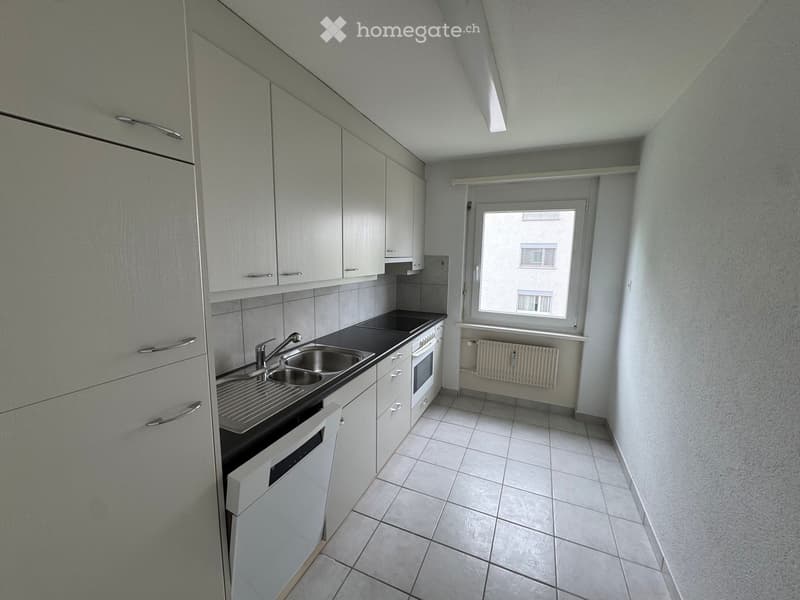 Modern kitchen with white cabinets, stainless steel sink, oven, and dishwasher. The space features tiled flooring and a window with a radiator below.