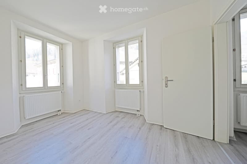 white walls, wooden floor, two large windows, two radiators, open door