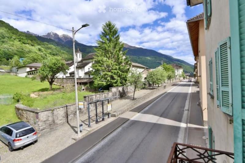 A quiet village street with a view of mountains, featuring a parked car, stone walls, and traditional houses.
