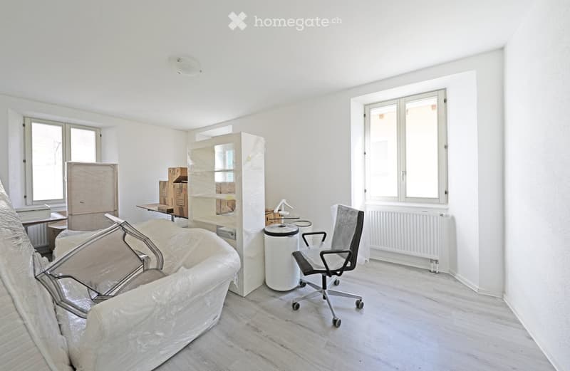 empty room, white walls, two windows, office chair, desk, boxes, wrapped furniture, trash bin, radiator
