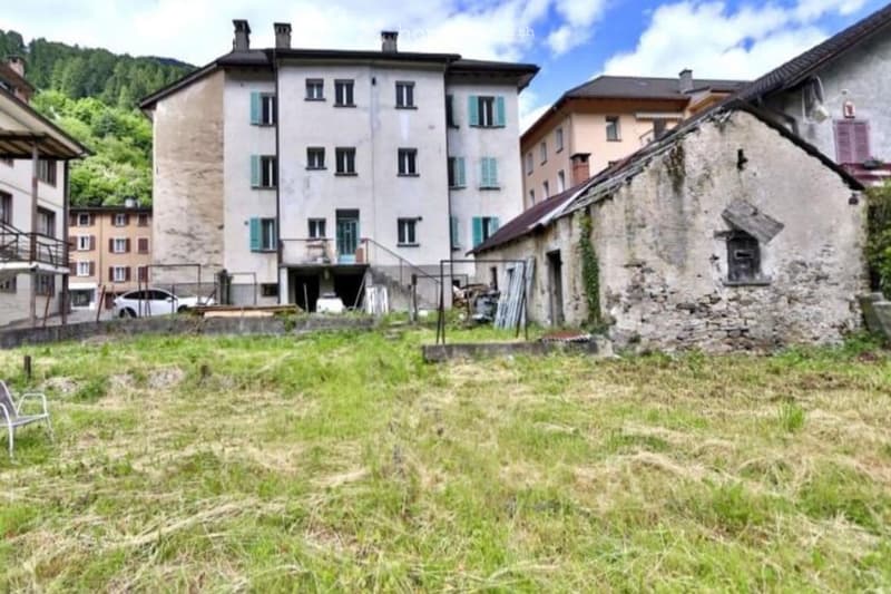 A multi-story building with multiple windows and shutters, situated in a rural area with a grassy yard and neighboring structures.