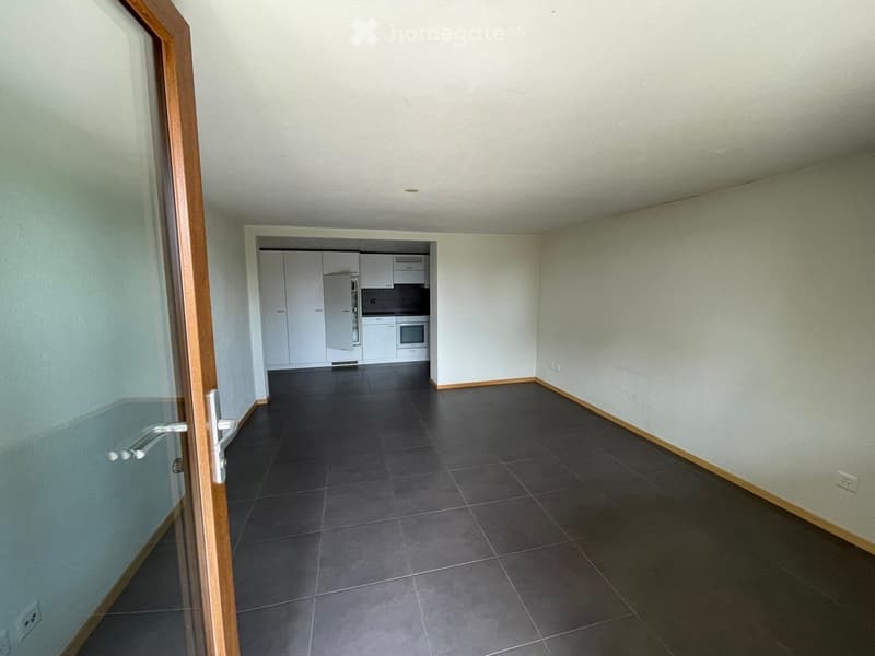 Empty room with tiled floor and white walls, featuring a doorway leading to a small kitchen area with white cabinets and appliances.