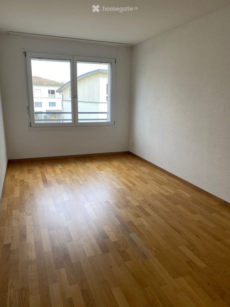 Empty room with wooden flooring and large windows, showing a view of neighboring buildings.