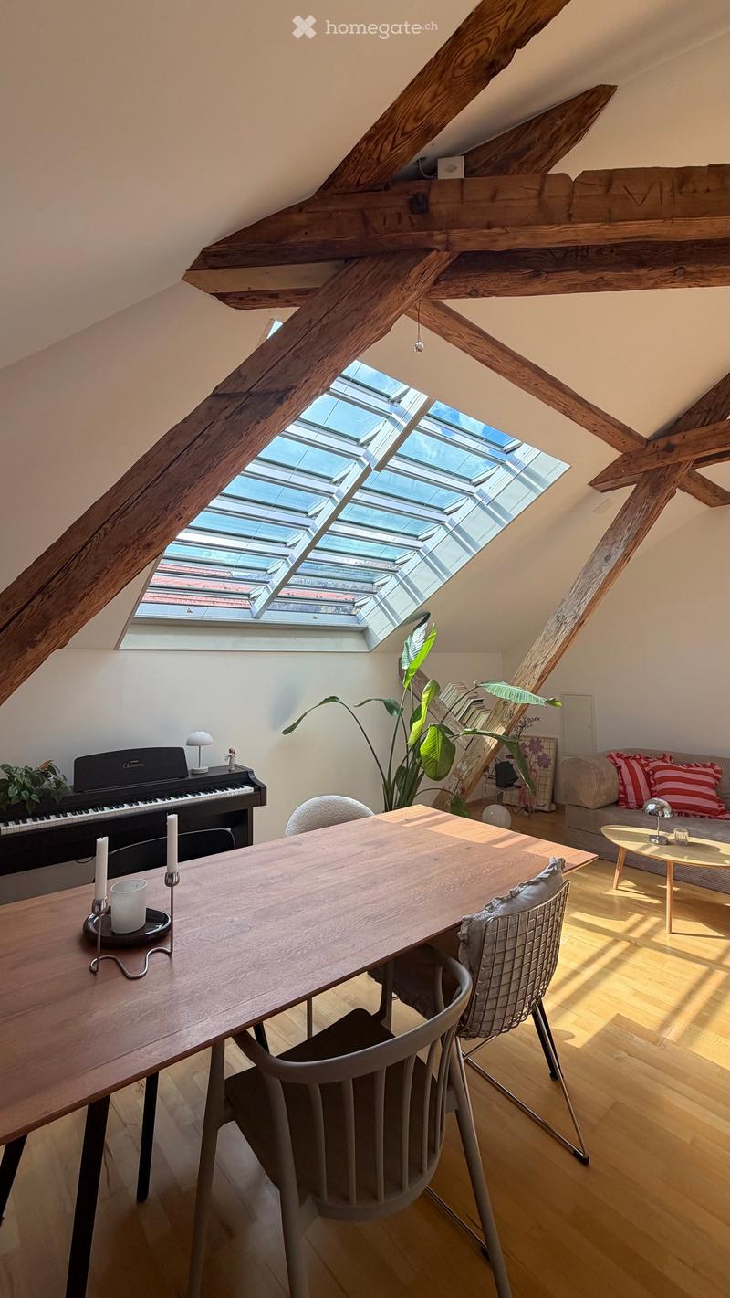 Bright, airy room with wooden beams and skylight windows, featuring a long wooden dining table, chairs, a sofa with striped cushions, a piano, and a potted plant.