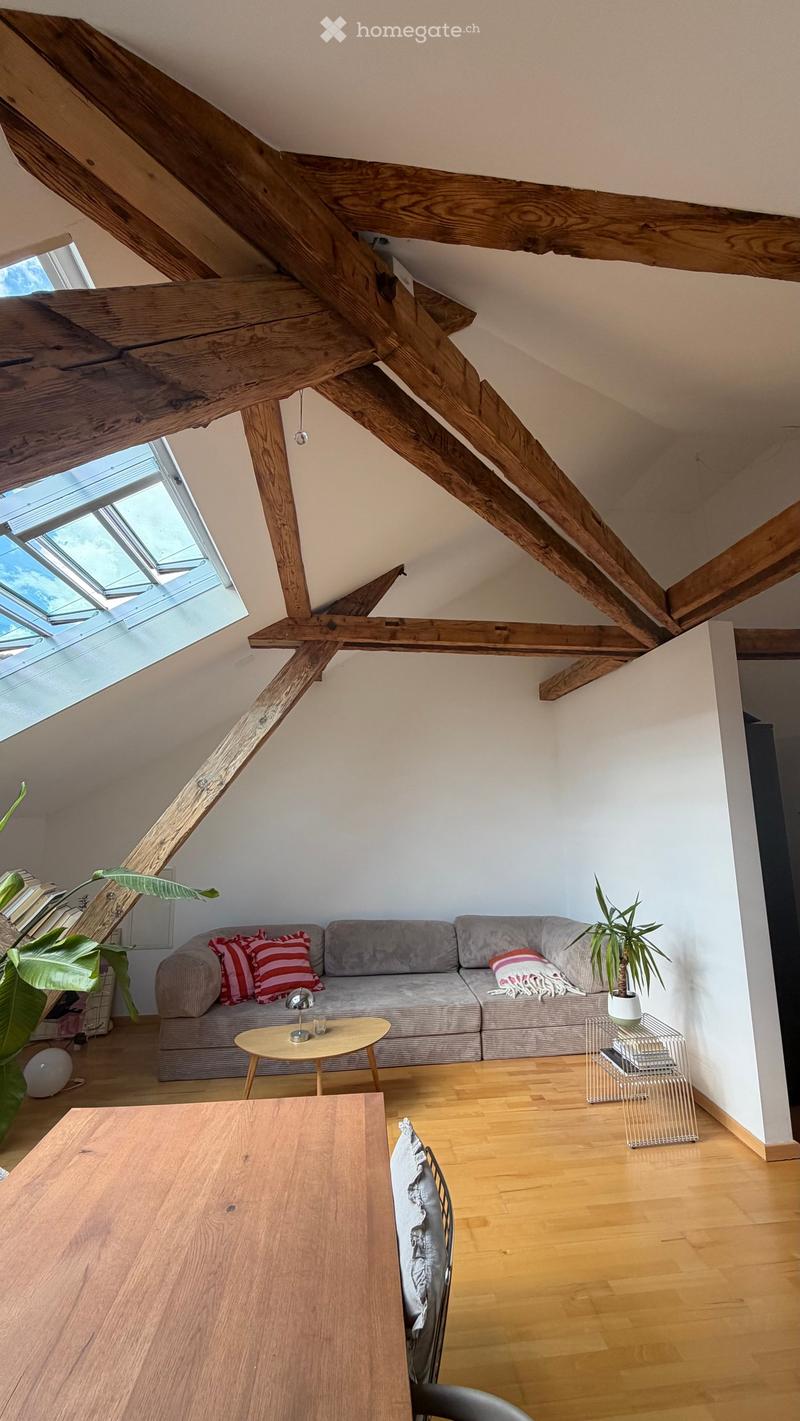 A modern living room with exposed wooden beams, a grey sofa with red and white pillows, a small wooden coffee table, a large wooden dining table, and plants. The room features wooden flooring and large skylights.