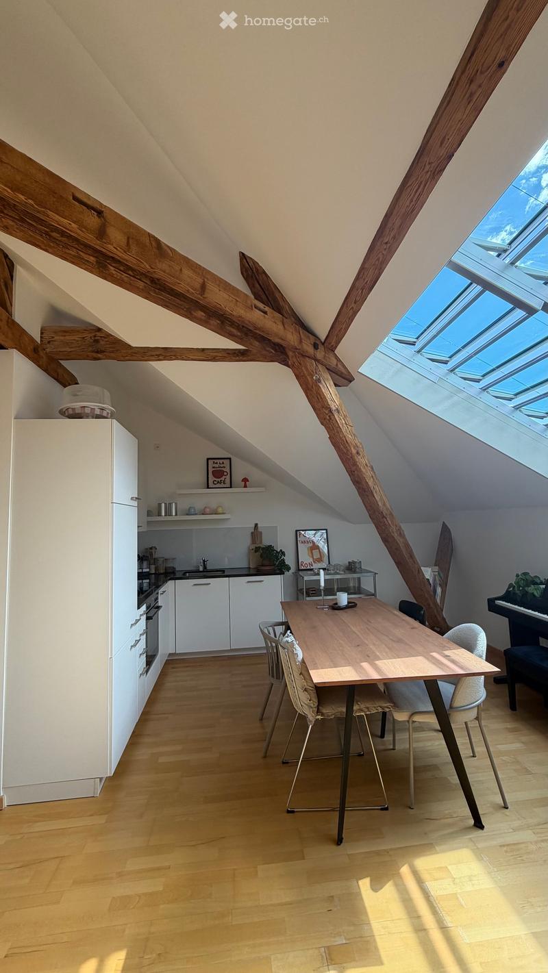 Bright, open loft-style room with wooden beams, skylights, a dining table with chairs, a modern kitchen area, and a piano in the corner.