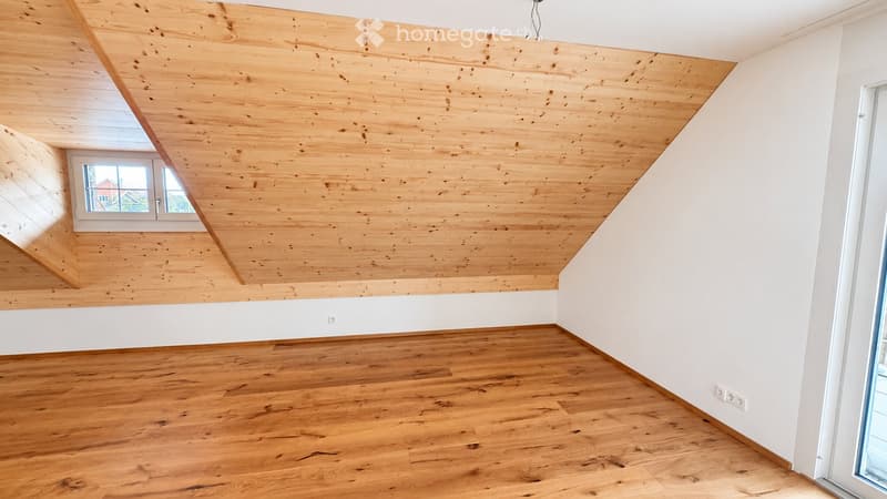 Empty room with wooden floor and slanted wooden ceiling, white walls, and a window letting in natural light.