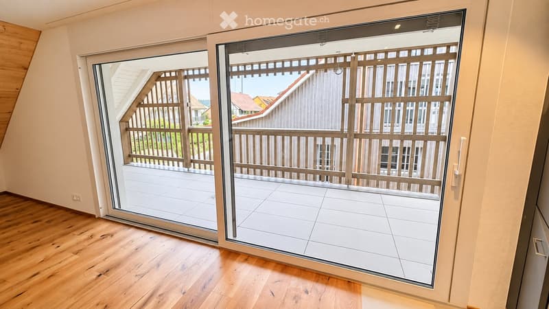 Sliding glass doors open to a tiled balcony with wooden railings, overlooking a residential area with houses and greenery.