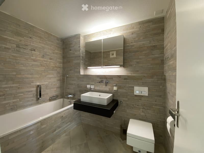 Modern bathroom with wooden tile walls, a bathtub, a floating sink, a toilet, and a large mirror with built-in lighting.