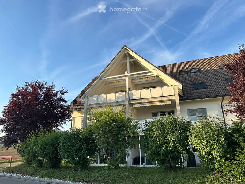 Two-story house with a pitched roof, balcony, and surrounding greenery.