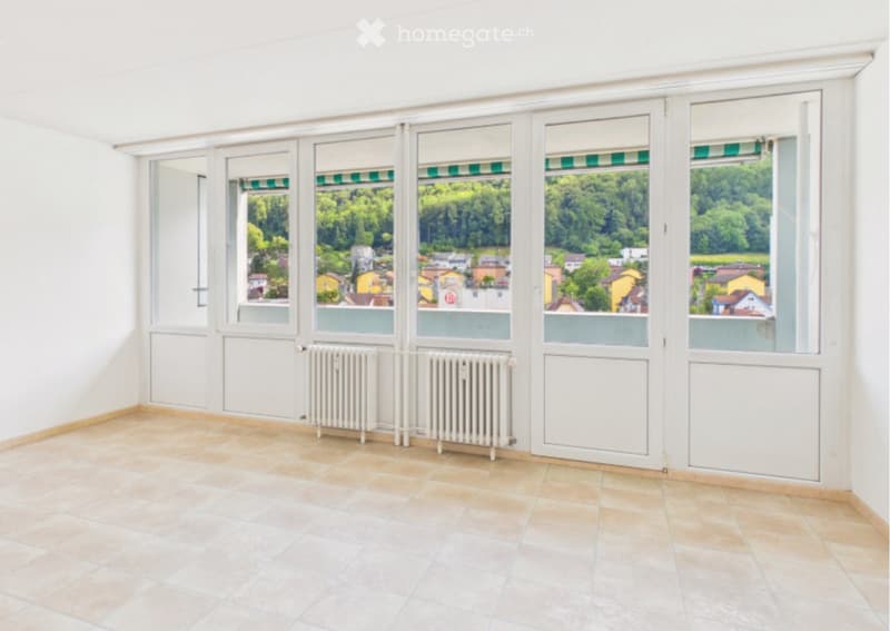 Empty room with tiled floor, large windows offering a view of a town and greenery, radiators, and white walls.