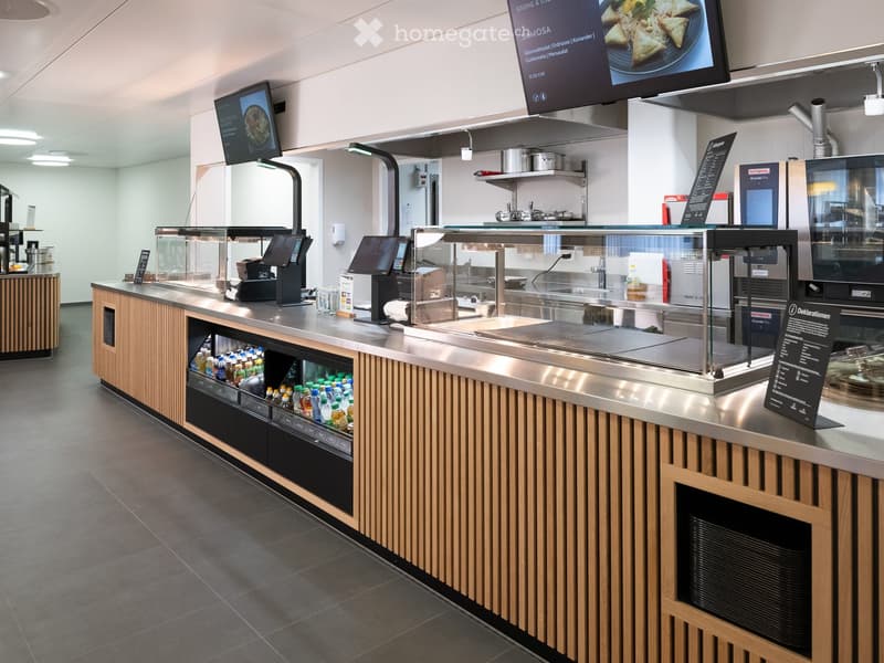 Modern kitchen area with stainless steel counter, digital screens, drinks dispenser, and various appliances.