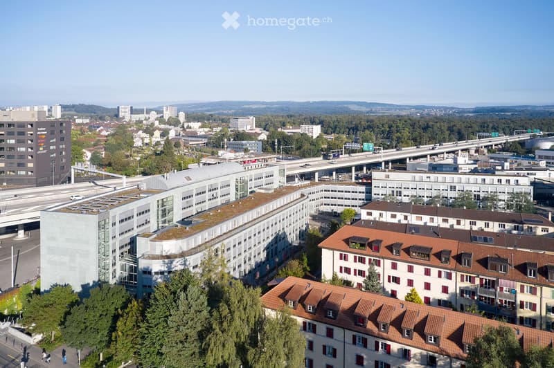 Aerial view of a cityscape with a mix of modern and traditional buildings, including a prominent modern building with a green roof, surrounded by trees and a highway.