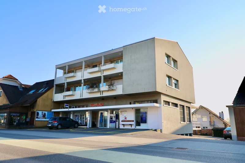 Modern multi-story building with balconies, a storefront labeled 'RAIFFEISEN', and a parking area with a car.