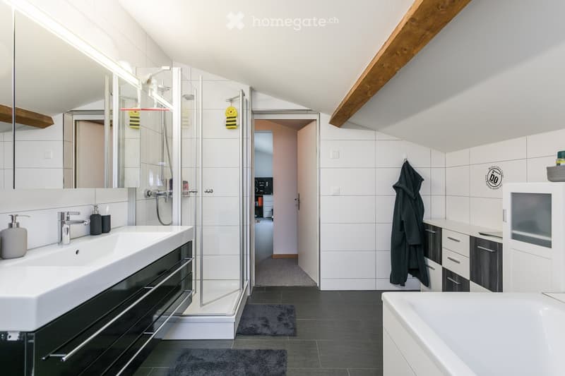 Modern bathroom with a large sink, bathtub, glass-enclosed shower, and white tiled walls. Features include a mirrored cabinet, a hanging bathrobe, and sleek black and white cabinetry.