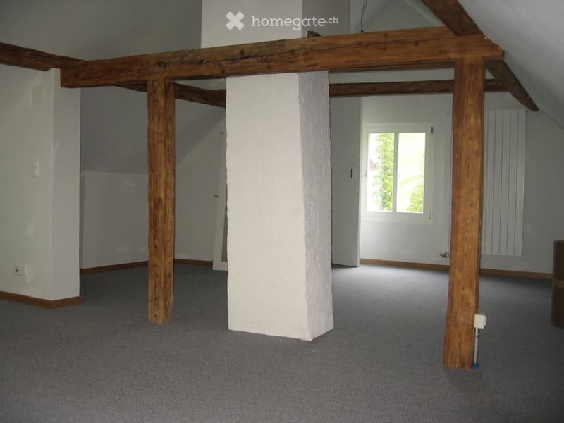 Empty attic space with exposed wooden beams and white plaster walls, featuring a window and carpeted floor.