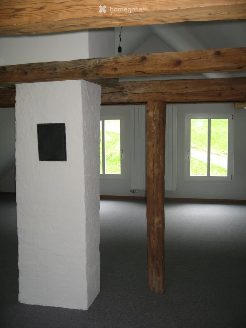 An unfurnished room with exposed wooden beams and a white pillar, featuring two windows with white frames and a grey carpeted floor.