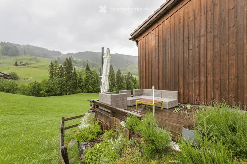 Wooden deck with outdoor furniture including a sofa, coffee table, and umbrella, surrounded by greenery and a scenic countryside view.