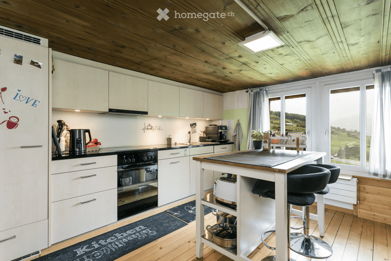 Modern kitchen with white cabinets, wooden ceiling, and a central island with bar stools. Appliances include a refrigerator, oven, and coffee maker. There is a view of a green landscape through large windows.
