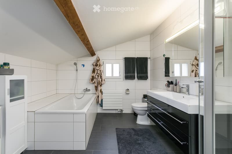 Modern bathroom with white tiles, a bathtub, toilet, double sink, and a large mirror. Features include a shower, towels, and a dark grey floor.