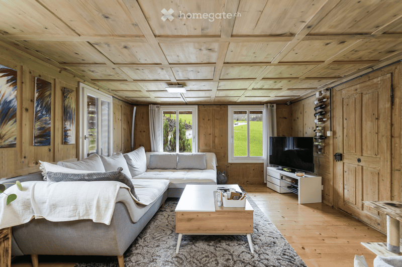 Cozy living room with wooden walls and ceiling, large windows, grey sectional sofa, coffee table, TV stand, and wine rack.