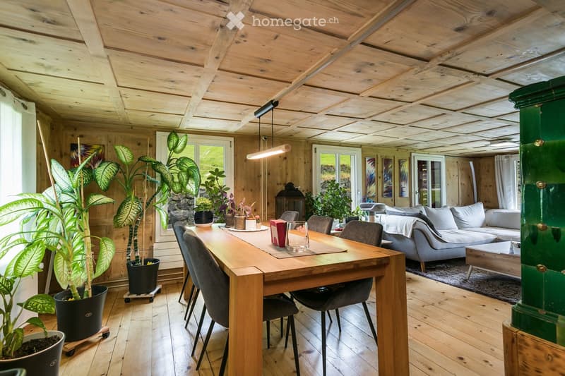 Bright, wooden-floored room with a dining area and living area. Features a long wooden dining table with chairs, a grey sofa, a coffee table, and several potted plants. The ceiling has exposed wooden beams and modern lighting.