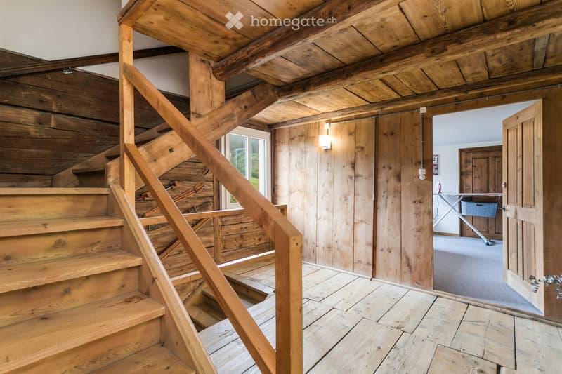 Wooden staircase with a handrail leads up, wooden walls and floor, visible doorway to another room with a washing machine and ironing board, natural lighting from a window.