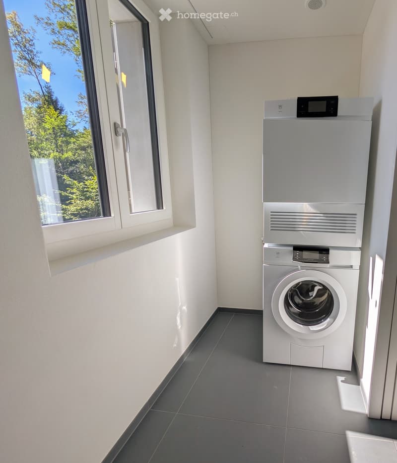 Modern, well-lit room with a washing machine and dryer unit, large window with a view of greenery.