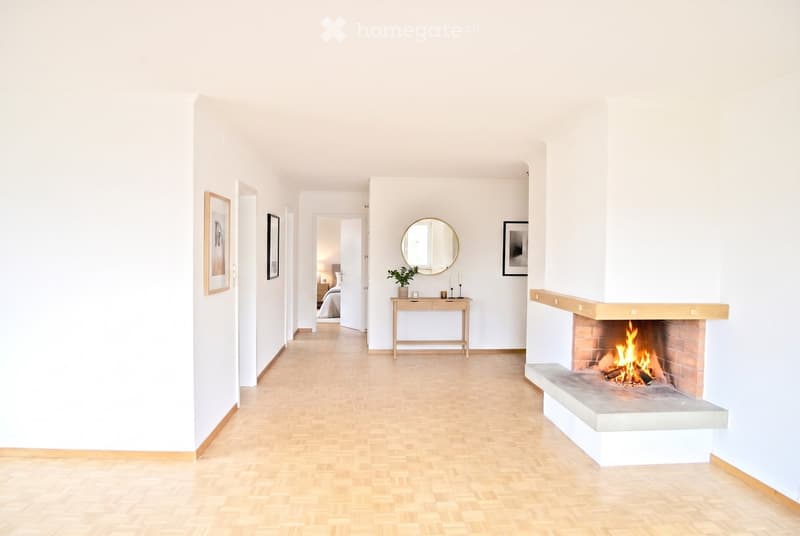 Bright hallway with wooden flooring, white walls, and a lit fireplace on the right side. A small table with a plant and candles is placed near the fireplace, and framed artwork adorns the walls. A bedroom is visible at the end of the hallway.