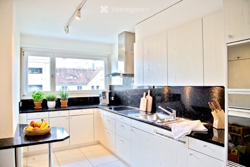 Modern kitchen with white cabinets, black countertops, stainless steel sink, and various appliances. Features include a knife block, coffee maker, potted plants, and a bowl of fruit on the counter.