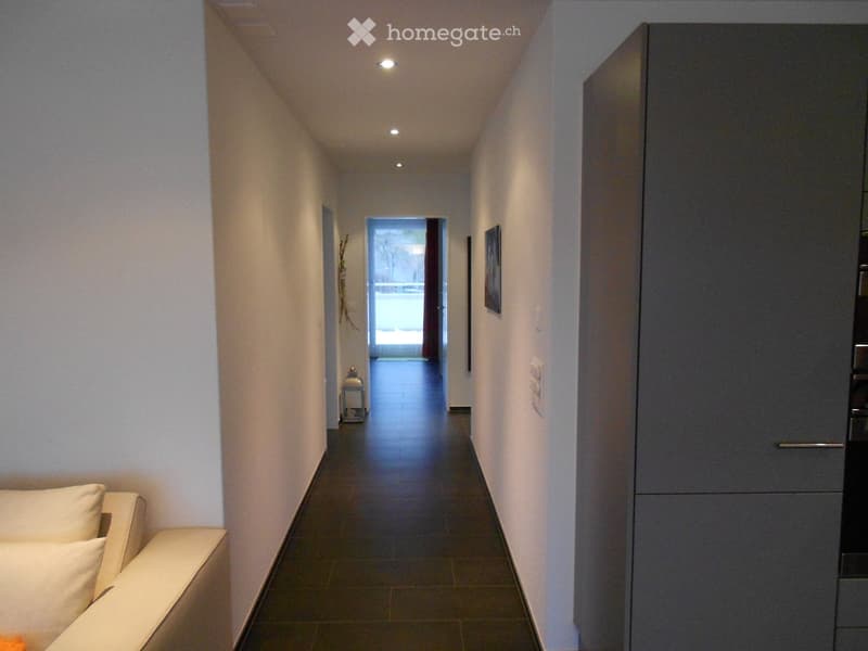 Modern hallway with white walls, dark tiled floor, recessed lighting, and a small bench with white cushions on the left side.