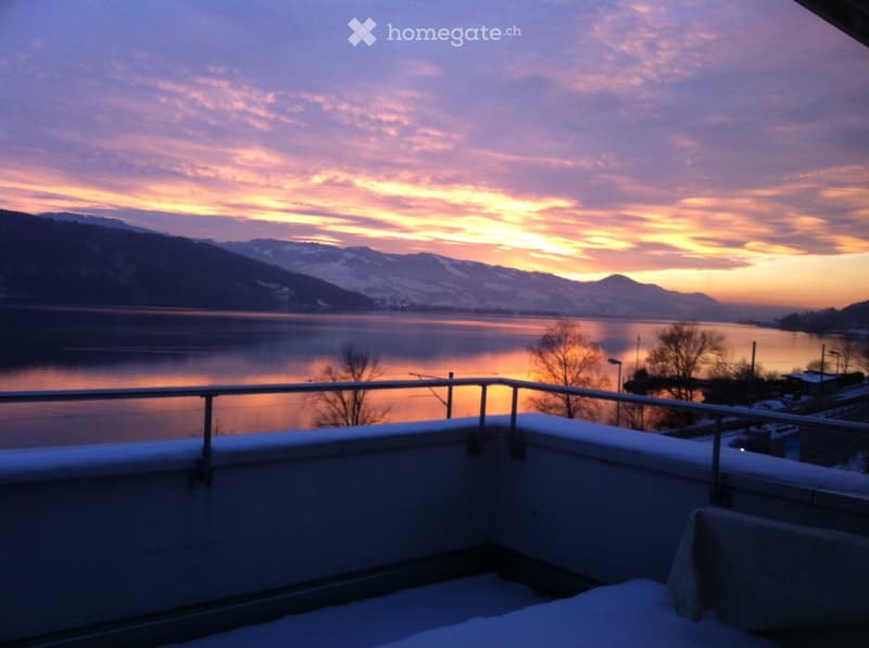 Balcony with snow-covered railing, overlooking a serene lake and snow-capped mountains during sunset.