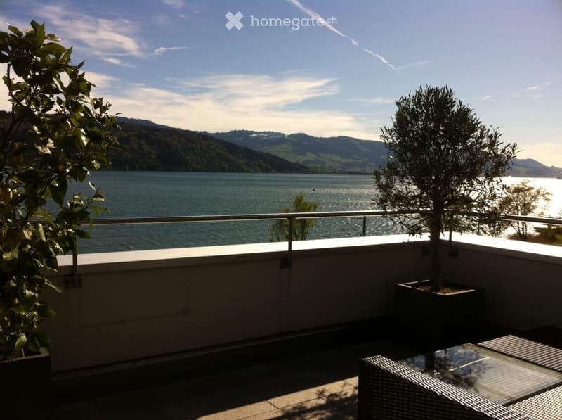 Balcony overlooking a lake with mountains in the background, featuring a glass table and wicker chairs.