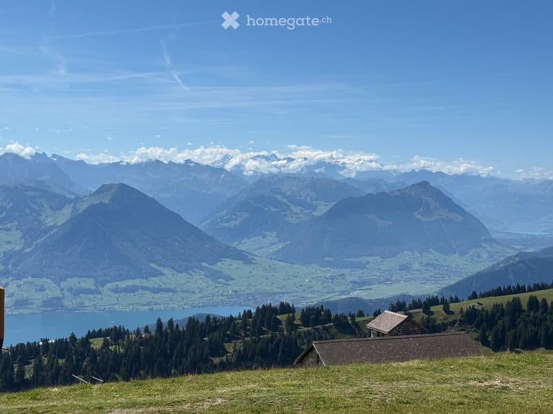 Panoramic view of a mountainous landscape with a lake in the foreground, surrounded by green fields and scattered trees.