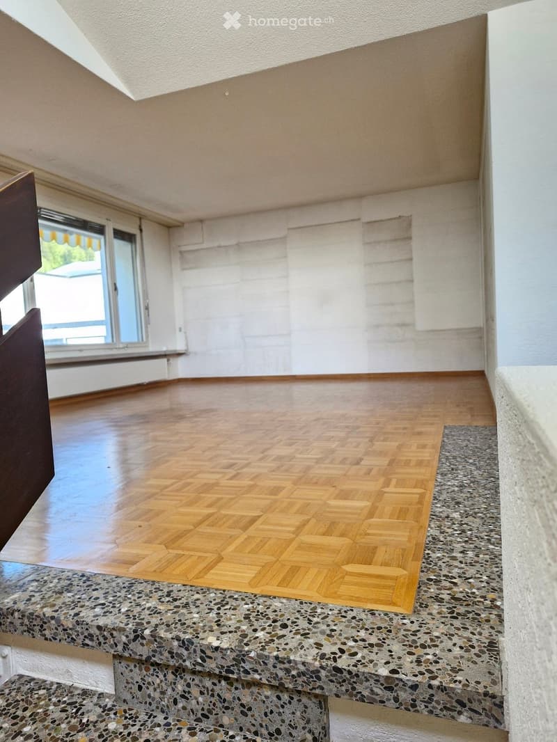 Empty room with wooden floor and large window, featuring a unique patterned wooden floor and a textured concrete step leading to the window area.