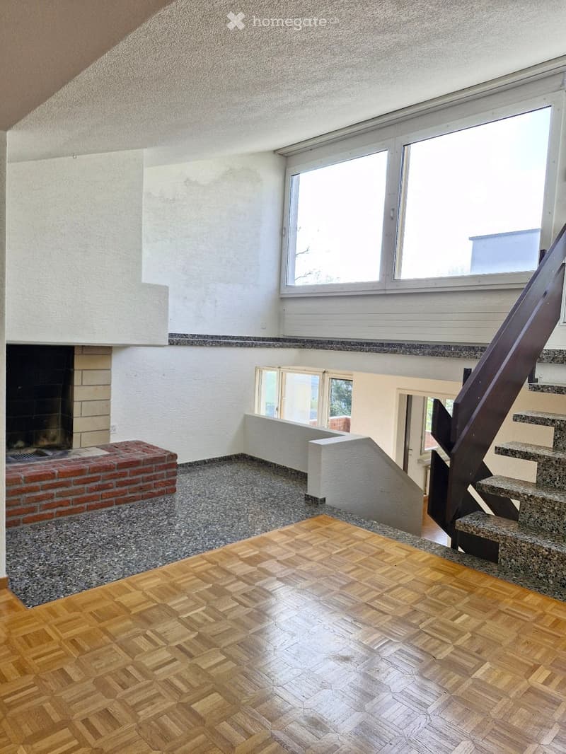 Modern living room with wooden herringbone flooring, fireplace, large windows, and a staircase leading upwards.