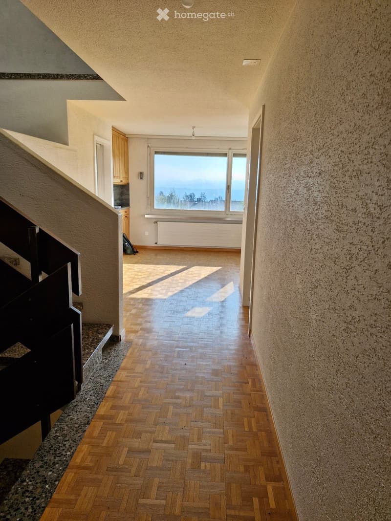 A long, narrow hallway with wooden herringbone flooring, textured walls, and a staircase on the left. A large window at the end offers a view of the sea.