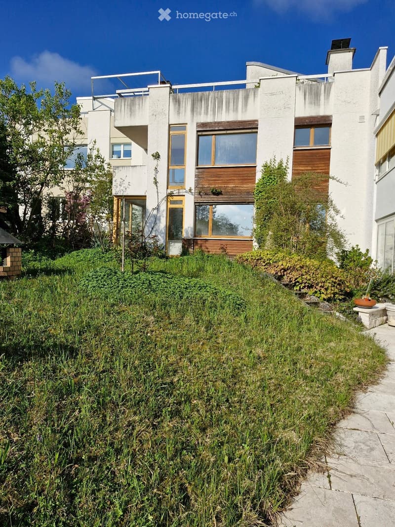 Modern two-story house with white walls, large windows, and a raised ground floor surrounded by greenery and plants.