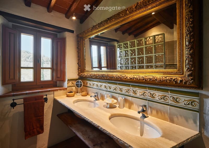 Elegant bathroom with double sinks, ornate mirror, decorative tiles, and wooden window shutters.