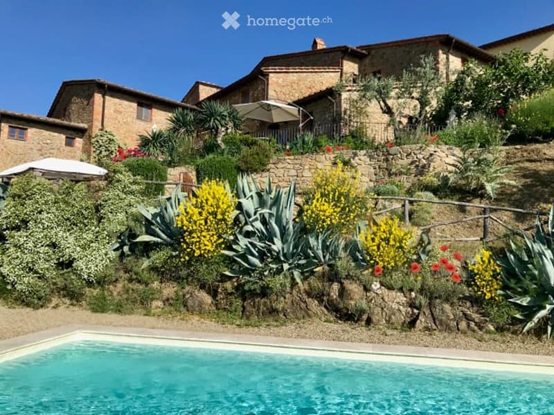 A scenic garden area with a swimming pool, surrounded by stone walls, colorful flowers, and lush greenery, with a rustic stone building in the background.